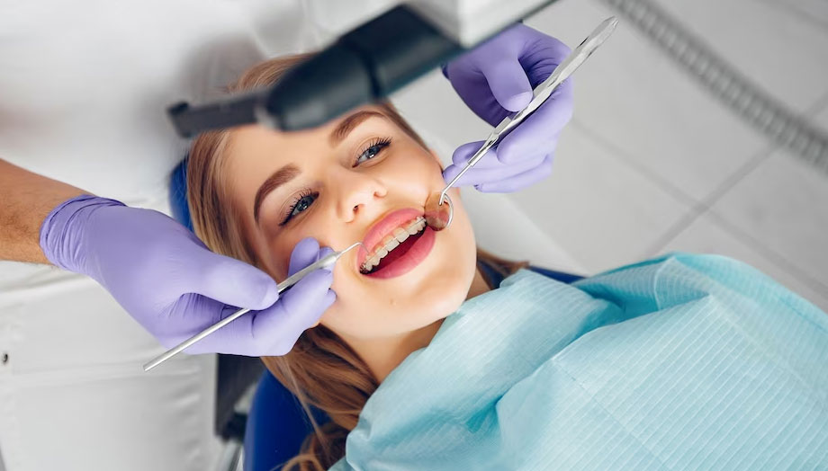 Imagen de una mujer sentada en una silla de dentista, sonriendo mientras un odontólogo la atiende. La mujer tiene la boca abierta y se puede ver que el odontólogo está examinando o tratando sus dientes. El odontólogo está usando guantes y tiene una herramienta dental en la mano. La mujer parece relajada y cómoda en el consultorio dental, y su sonrisa sugiere que se siente segura y en buenas manos. La imagen transmite confianza y profesionalismo en su tratamiento de diseño de sonrisa sin brackets